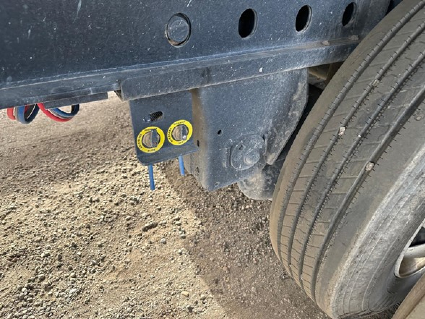 Trailer hitch area showing two yellow-capped ports and a blue-handled bolt under a dark blue chassis near a tire, with round and rectangular holes in the frame.