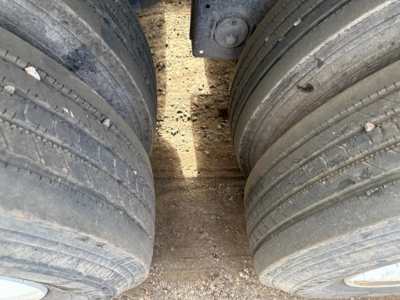 Flatbed trailer wheels and dual tires viewed from between tires, dirt/grime on ground, part of undercarriage visible.