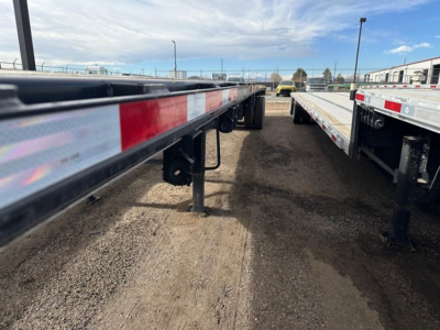 Trailer frame side with reflective red/white tape and support legs, long flatbed trailer in a lot, other trailers in background.
