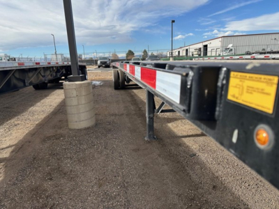 Flatbed trailer side rail with reflective red-white tape and yellow warning label, level ground with gravel, warehouse buildings in background.