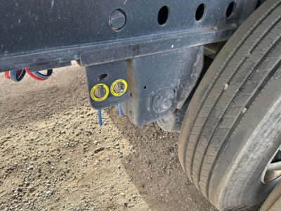 Trailer hitch area showing two yellow-capped ports and a blue-handled bolt under a dark blue chassis near a tire, with round and rectangular holes in the frame.