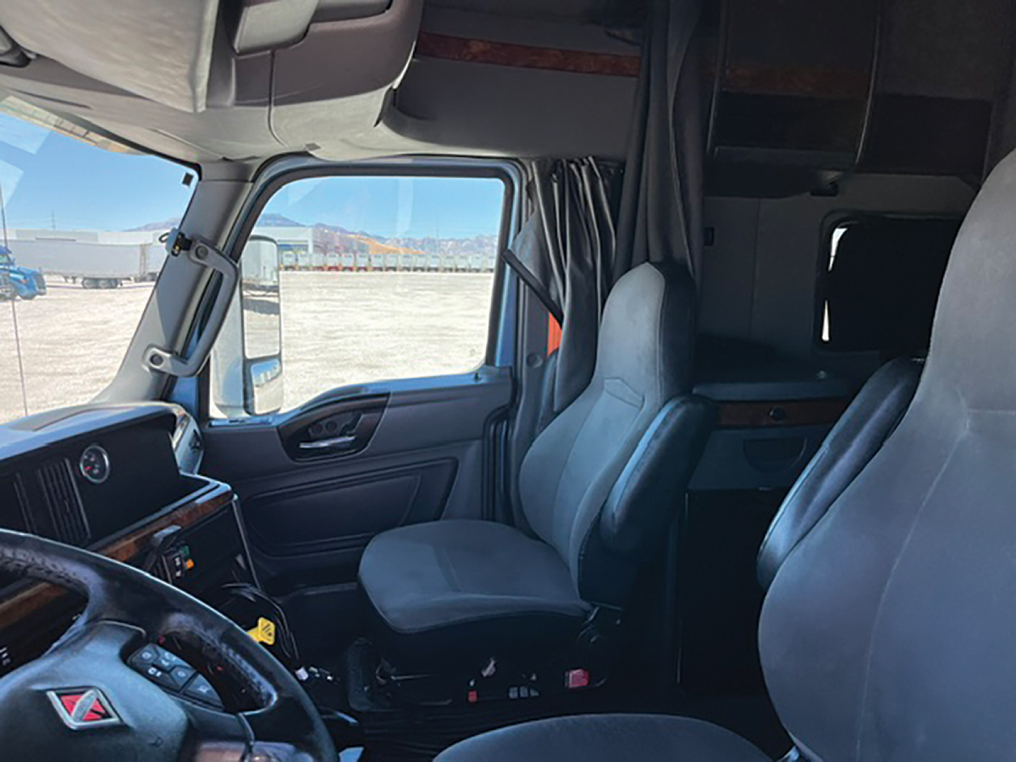 Interior of a 2019 International LT625 truck cab with two front seats, dashboard, steering wheel, and a sleeping area behind. Cab view shows side window, door, and curtain.