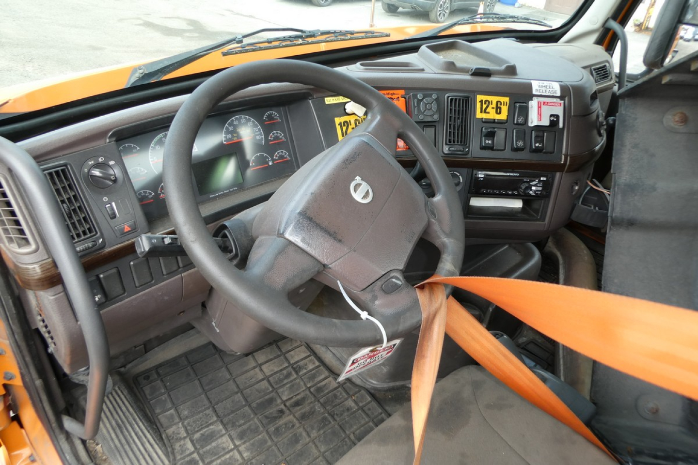 Volvo VNM42T truck interior showing steering wheel, dashboard gauges, and seat belt, with yellow warning labels on dash.
