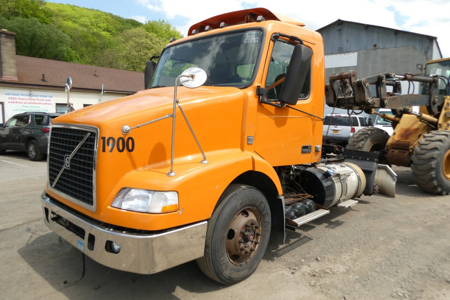 Orange Volvo trucks cab with number 1900 on hood, side mirrors, fuel tanks, and exposed engine bay; daycab configuration in yard.
