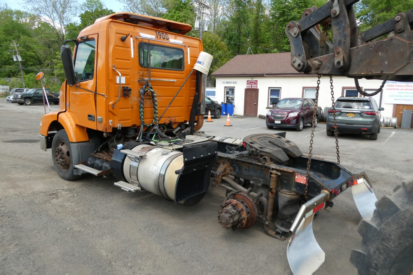 Orange Volvo VNM42T truck cab with disassembled front axle and frame sections in a lot, visible fuel tank and machinery around.