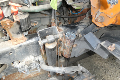 Vehicle chassis with exposed hydraulic lines and rusted brackets; orange component on right; metal frame with weathered hoses and fittings visible.