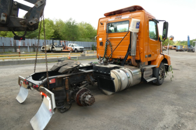 Volvo VNM42T conventional daycab truck chassis with fifth wheel and fuel tank, orange cab, detached rear underride guard and components.