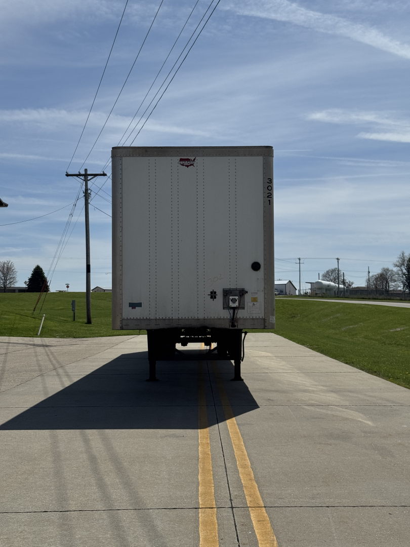 Trailer rear view showing plain white Wabash Duraplate box with rear doors, standing on two support legs on a road, with license-free area and power lines in background.