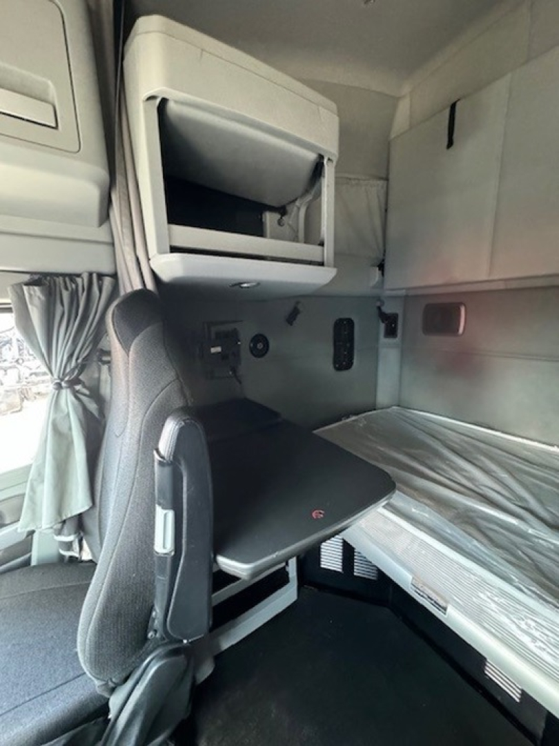 Cab interior of a 2022 Kenworth T680 sleeper. Image shows a compact bunk area with a fold-down desk/table, overhead storage compartment, and window curtain on left.