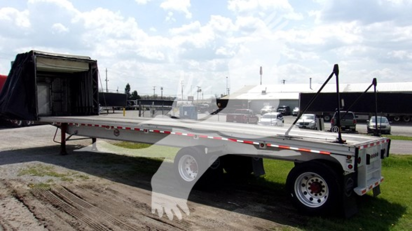 Curtainside trailer with open ramp showing aluminum flatbed and tandem axles, loading area visible under a blue sky.