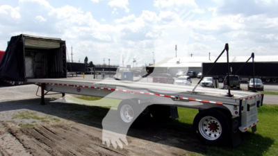 Curtainside trailer with open ramp showing aluminum flatbed and tandem axles, loading area visible under a blue sky.