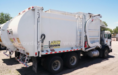 Peterbilt 320 garbage truck rear-side view showing large aluminum waste body with 'Slow Down' decal and triple rear axles.