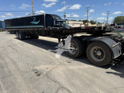 Flatbed trailer with a black Fontaine XTP 4880SS2 long trailer on a lot. Showcases rear tandem axles, side rails, and black cab with chrome mirrors.