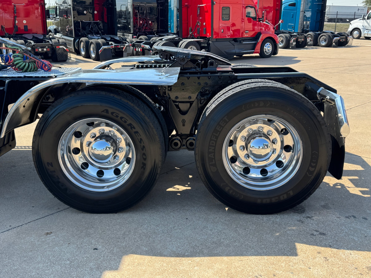 Dual rear wheels of a Kenworth W900L tractor showing chrome hubcaps, black chassis, and chrome fenders with a truck yard in background.