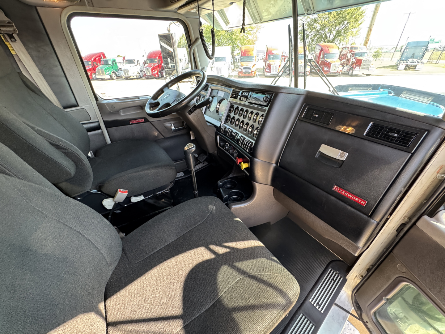 Kenworth W900L interior cab showing steering wheel, dashboard with control switches, gear lever, and seating. Visible text includes windshield and dash area but no legible model details beyond general cab layout.