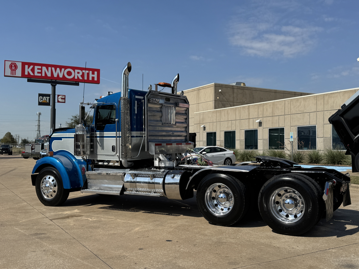 Kenworth W900L truck in a dealership lot showing chrome exhaust stacks and dual rear axles.
