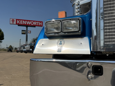 Kenworth W900L front bumper and headlight assembly with chrome grille and Kenworth sign in background.
