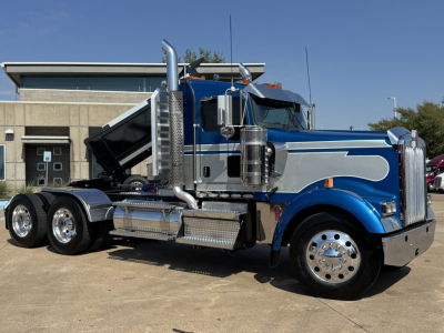 Kenworth W900L conventional daycab truck with a blue and silver paint scheme, chrome exhaust stacks, polished wheels, and a raised dump bed showing the truck's side profile.