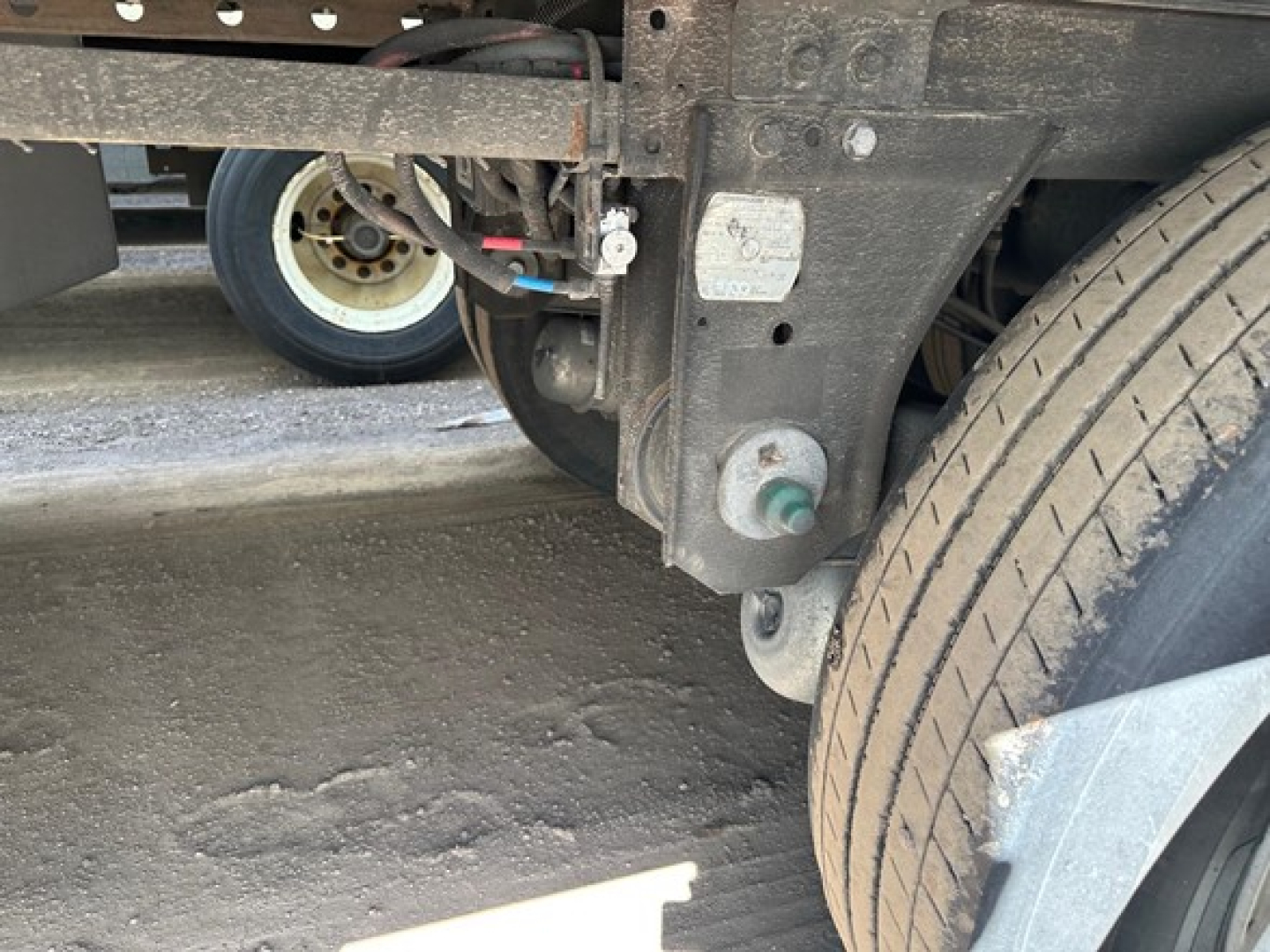 Trailer chassis with rear wheel and suspension; visible labels and components undercarriage; dirt and road grime on frame; small white/green caps and wiring harnesses.