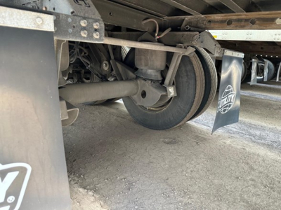 Trailer undercarriage showing axle, suspension, and wheel. Visible components include a round wheel hub, leaf spring, brake drum, and mud flaps with logos.