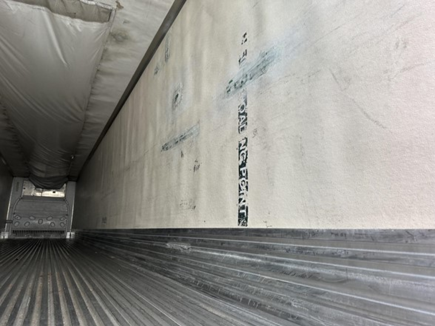 Trailer interior with ribbed metal floor and sidewall insulation, showing a reefer unit interior and rear stair area edge; faint vertical lettering on side wall.