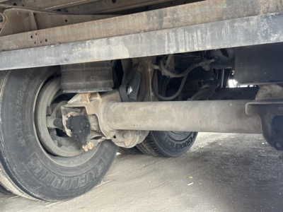 Trailer undercarriage showing wheel with tire, suspension axle, and trailer frame. Visible components include leaf spring, driveshaft area, and hydraulic lines.