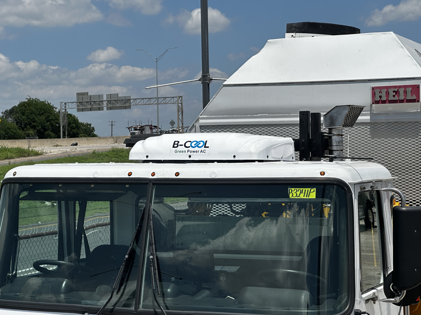 Truck cab with B-Cool Green Power AC rooftop unit and windshield details visible; small yellow stock number tag on windshield; white cab and side mirror; part of a refrigerated box behind.