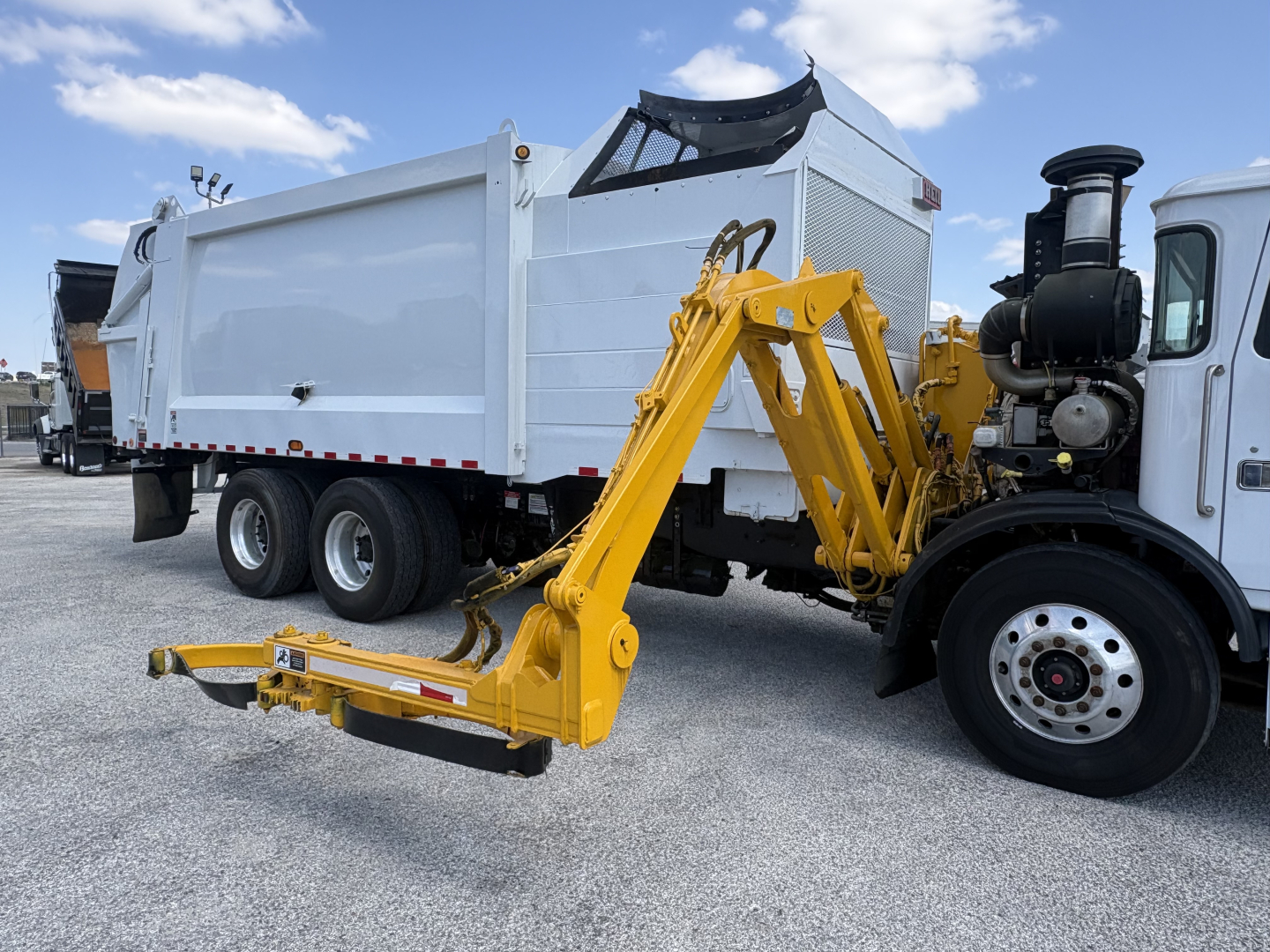 Autocar XPEDITOR truck with front attachment derrick and dump body, side view showing white cab, black engine area, and yellow hydraulic arm in a loading yard.