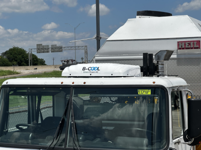 Truck cab with B-Cool Green Power AC rooftop unit and windshield details visible; small yellow stock number tag on windshield; white cab and side mirror; part of a refrigerated box behind.
