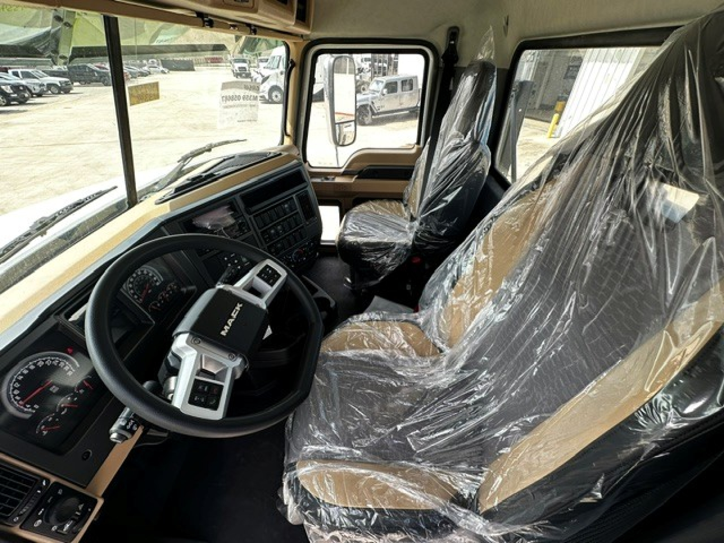 Cab interior of new 2027 Mack GR64F showing tan and black seats wrapped in plastic, steering wheel, dashboard, and large windows.