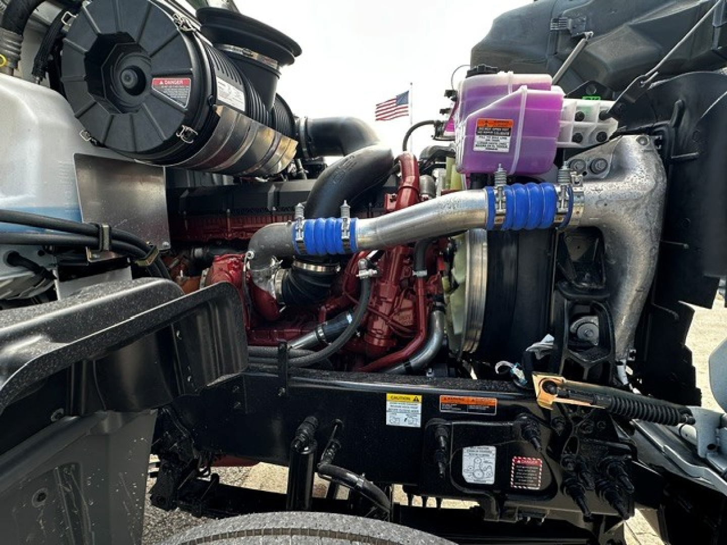 Truck engine bay of 2027 Mack GR64F showing radiator, coolant reservoir, hoses, turbocharger, and engine components with purple coolant reservoir and blue hose clamps.