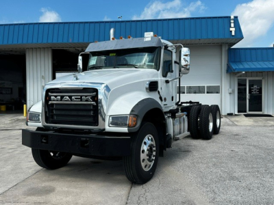 Mack GR64F cab and chassis truck in white parked outside a dealership. Prominent front grille with MACK lettering, large bumper, dual rear axles, side mirrors, and steps.