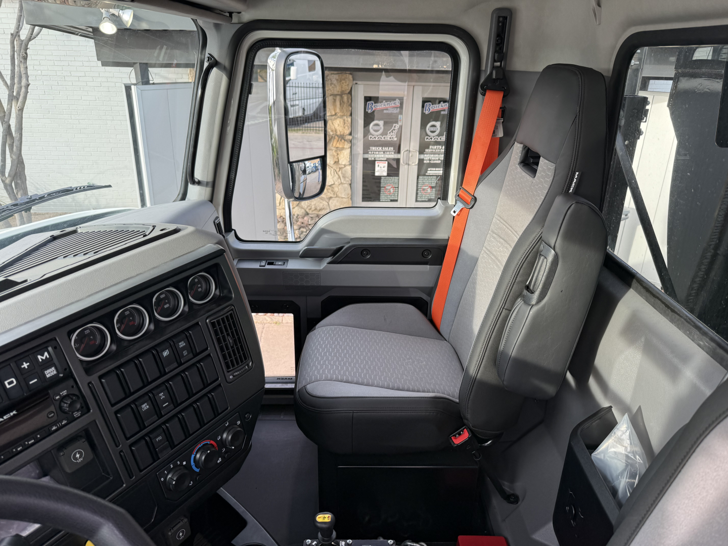 Truck cab interior of a 2026 Mack GR64F showing driver seat and dashboard with steering wheel, multiple gauges, climate controls, orange seat belt, and window view.