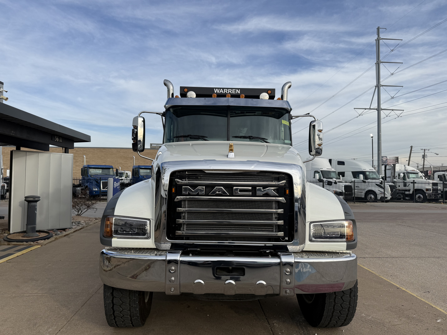 Mack dump truck front view showing grille with large MACK lettering, bumper, headlights, and towing hooks; windshield with side mirrors and a roof marker light.”