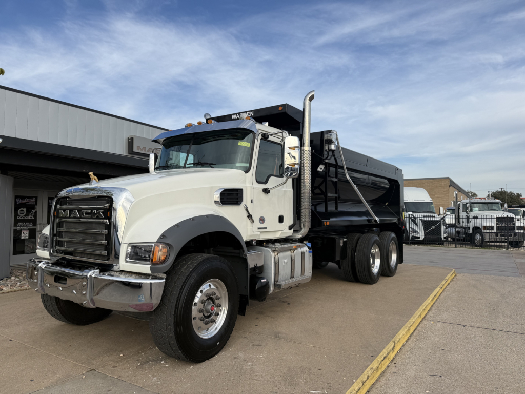 Dump truck in showroom setting showing front and left side, Mack grille, GR64F model, white cab, black dump bed, heavy-duty tires, chrome bumper and steps.