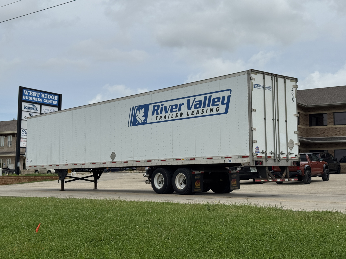 Utility semi-trailer in a dealership lot showing River Valley Trailer Leasing logo, tandem axles, white box trailer, rear doors, and a pickup truck attached.