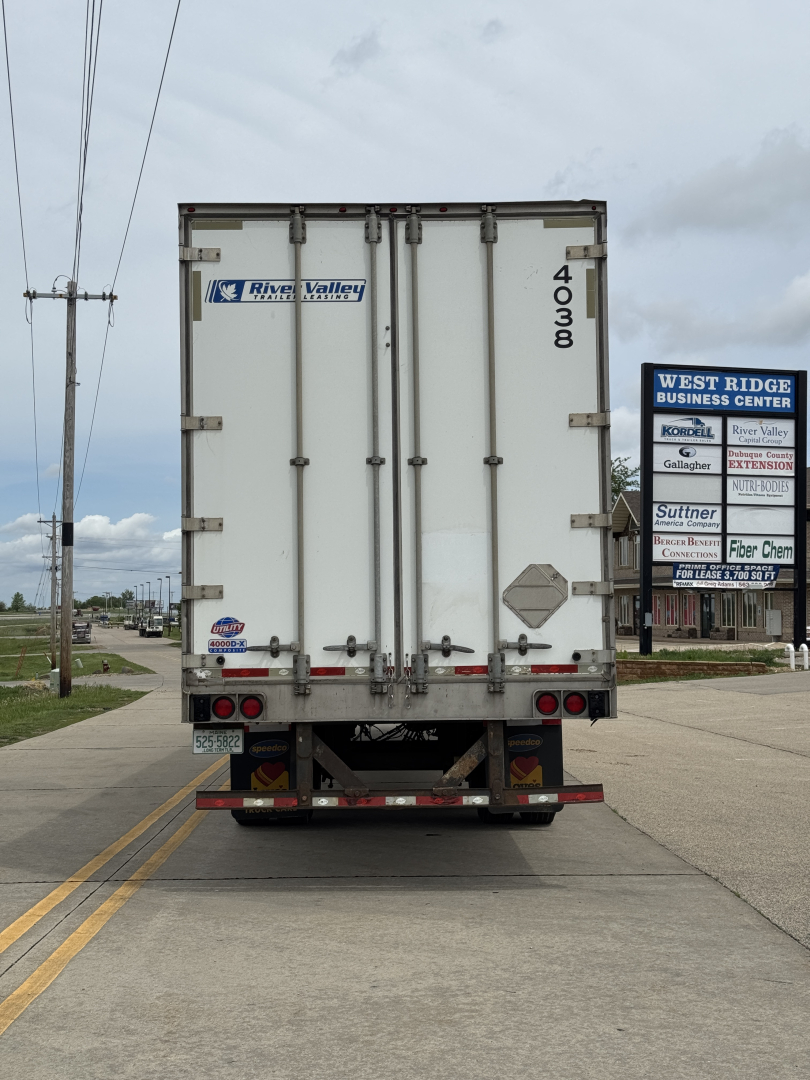 Rear of a white cargo trailer with River Valley branding, doors closed, model number 4003 or 4000 on the right door, taillights and bumper visible, street and signs in background.