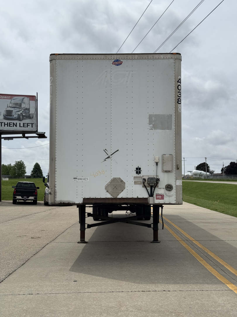 Utility 4000 Dx trailer rear view showing raised legs, rear doors, bumper with lights, and stickers/markings on a paved lot.