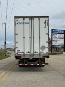 Rear of a white cargo trailer with River Valley branding, doors closed, model number 4003 or 4000 on the right door, taillights and bumper visible, street and signs in background.