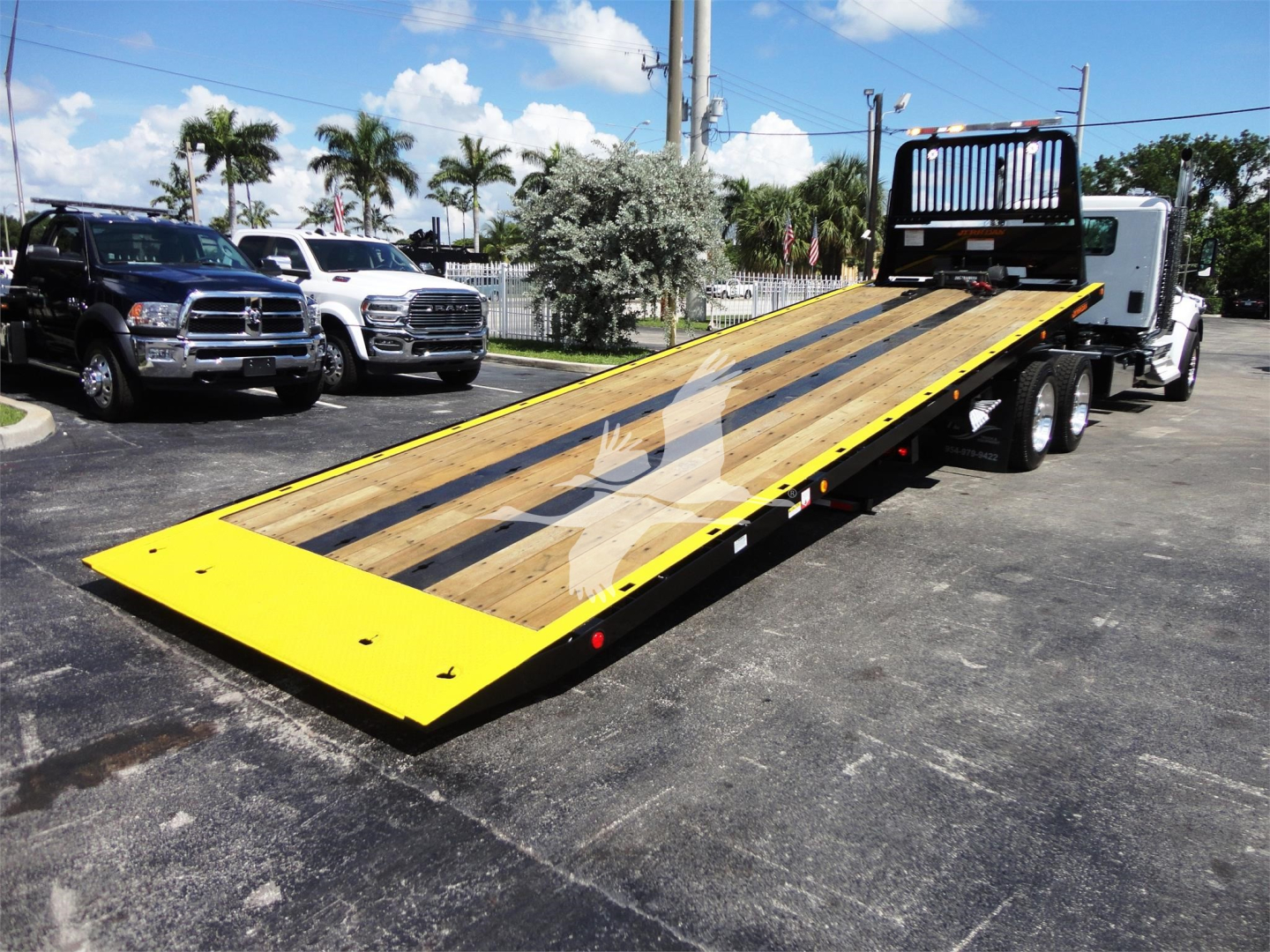 Kenworth W990 rollback truck with a long wooden flatbed and yellow edges, parked on pavement with other trucks nearby.