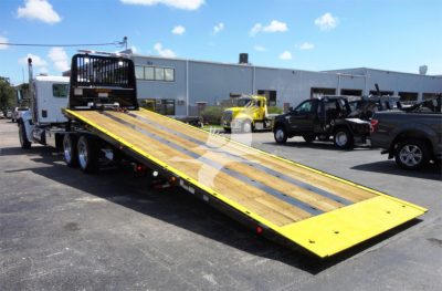 Kenworth W990 truck on a rollback with a long yellow tilt bed extended, in a dealership lot with other trucks and industrial buildings in background.