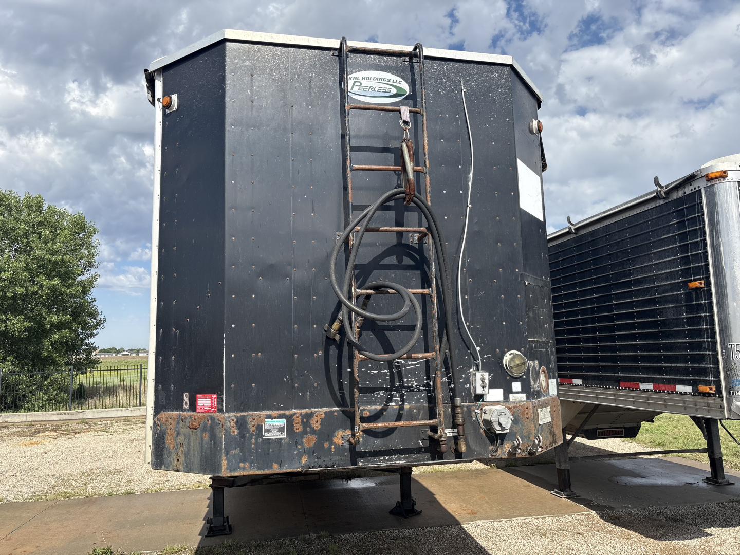 Rear view of a black trailer with a coiled hose on a rack, bumper guards, and registration stickers; visible are support legs and a steel frame, with weathered surfaces.