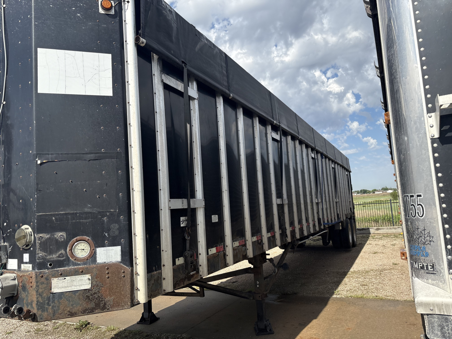 Trailer side view showing a black dump-style apron hopper trailer with support legs down and reflective tape along the frame. Weathered metal with rivets and a white rectangular label on the front corner.