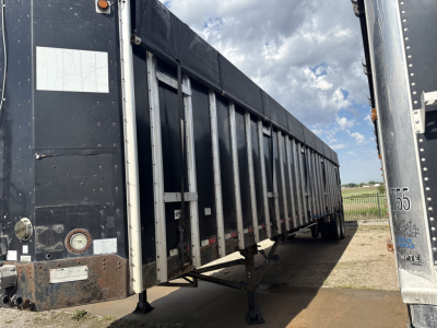 Trailer side view showing a black dump-style apron hopper trailer with support legs down and reflective tape along the frame. Weathered metal with rivets and a white rectangular label on the front corner.