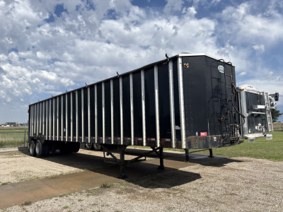 Trailer in-service showing black steel-sided trailer with dual rear axles and gooseneck hitch, side rails, and landing gear.