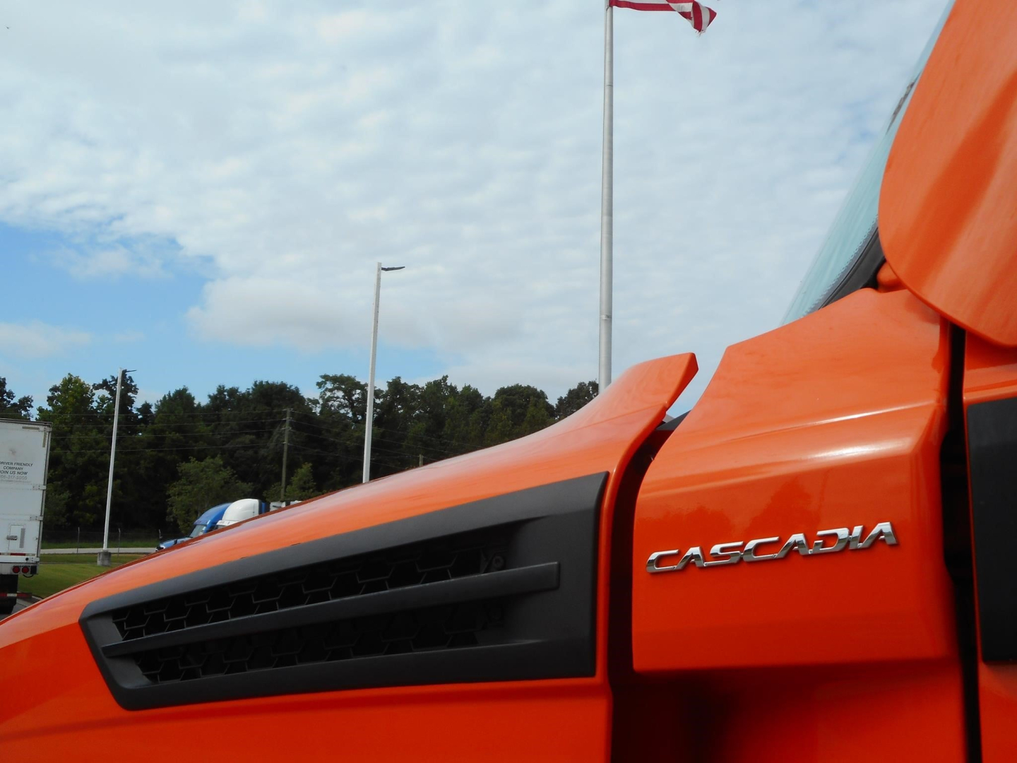 Orange Freightliner Cascadia truck cabin with 'CASCADIA' badge visible, side air intake vent and blue sky background.