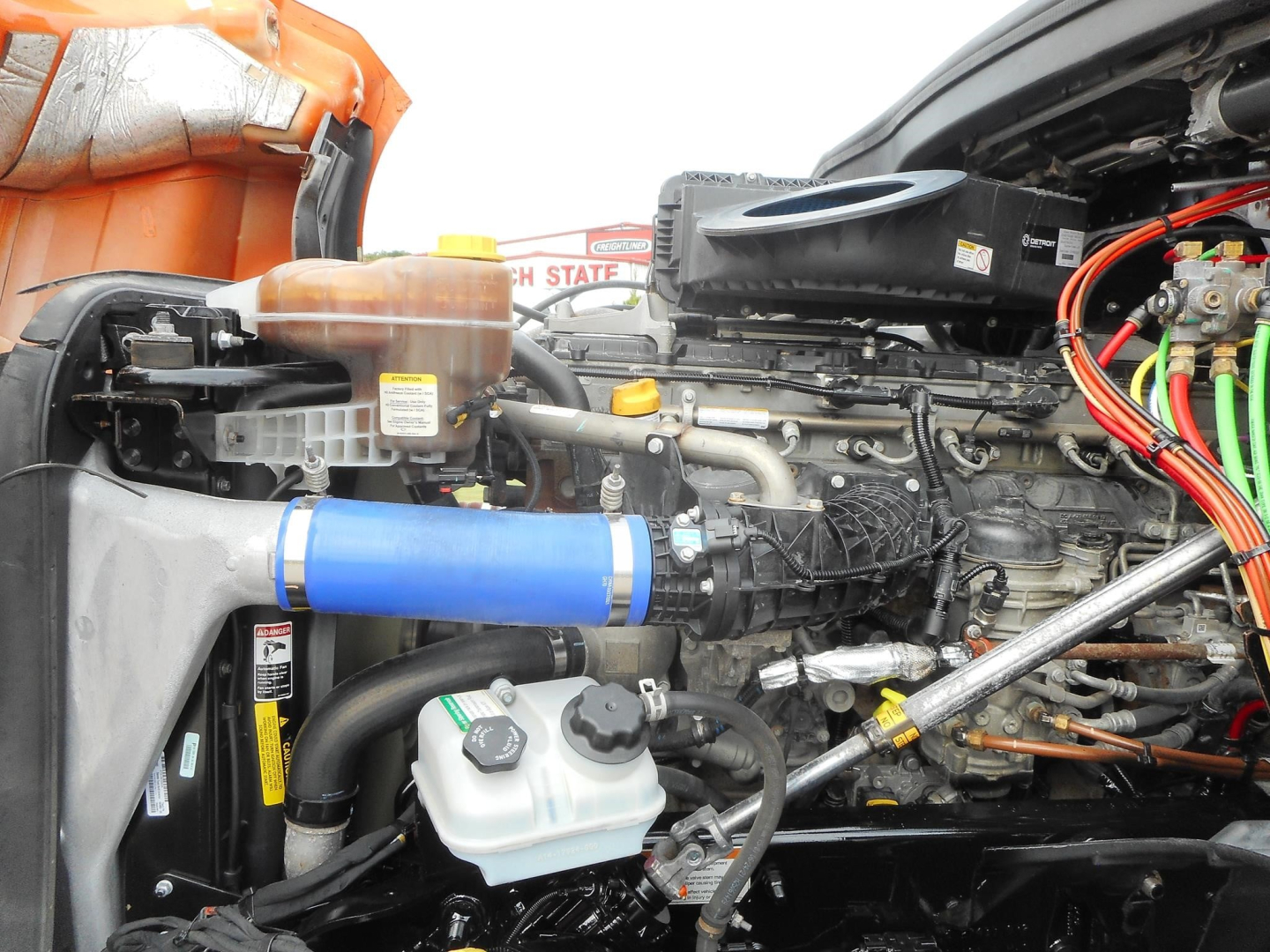 Freightliner Cascadia engine bay showing blue intercooler pipe, coolant reservoir, and various hoses and wiring in-view.