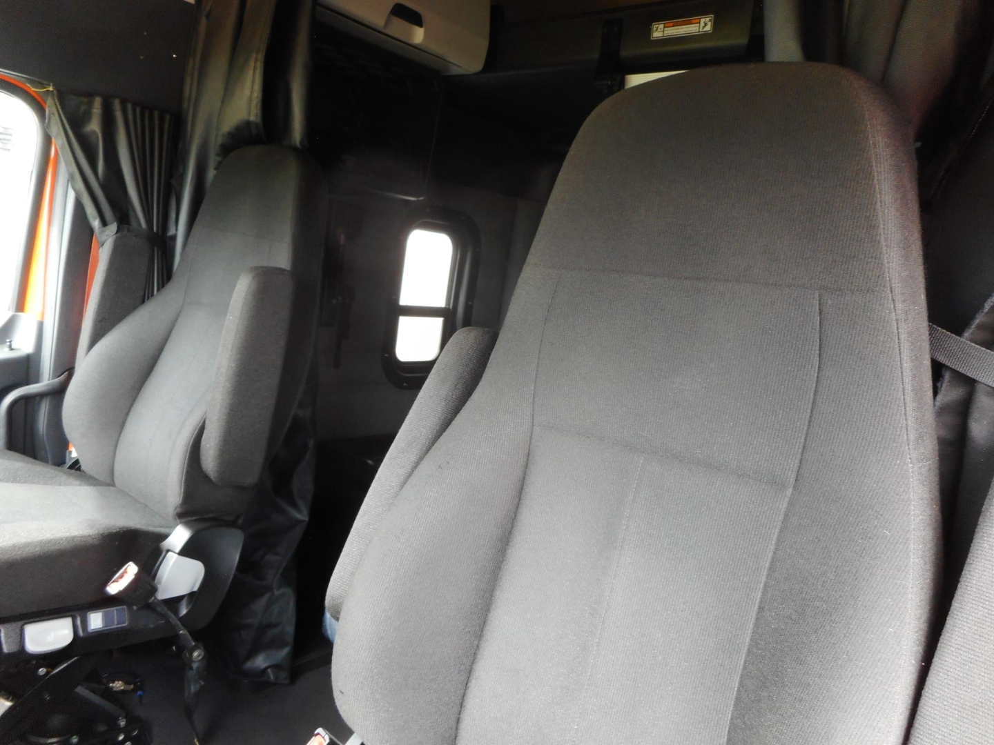 Cab interior of a Freightliner Cascadia showing driver and passenger seats and a small window; dark fabric seats.
