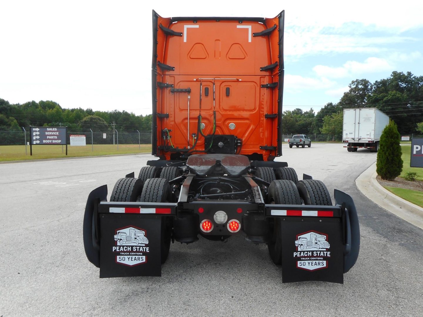 Rear view of a Freightliner Cascadia truck tractor with bright orange sleeper cab, dual rear axles, mud flaps with Peach State 50 Years logos, and view of fifth wheel and coupling area.