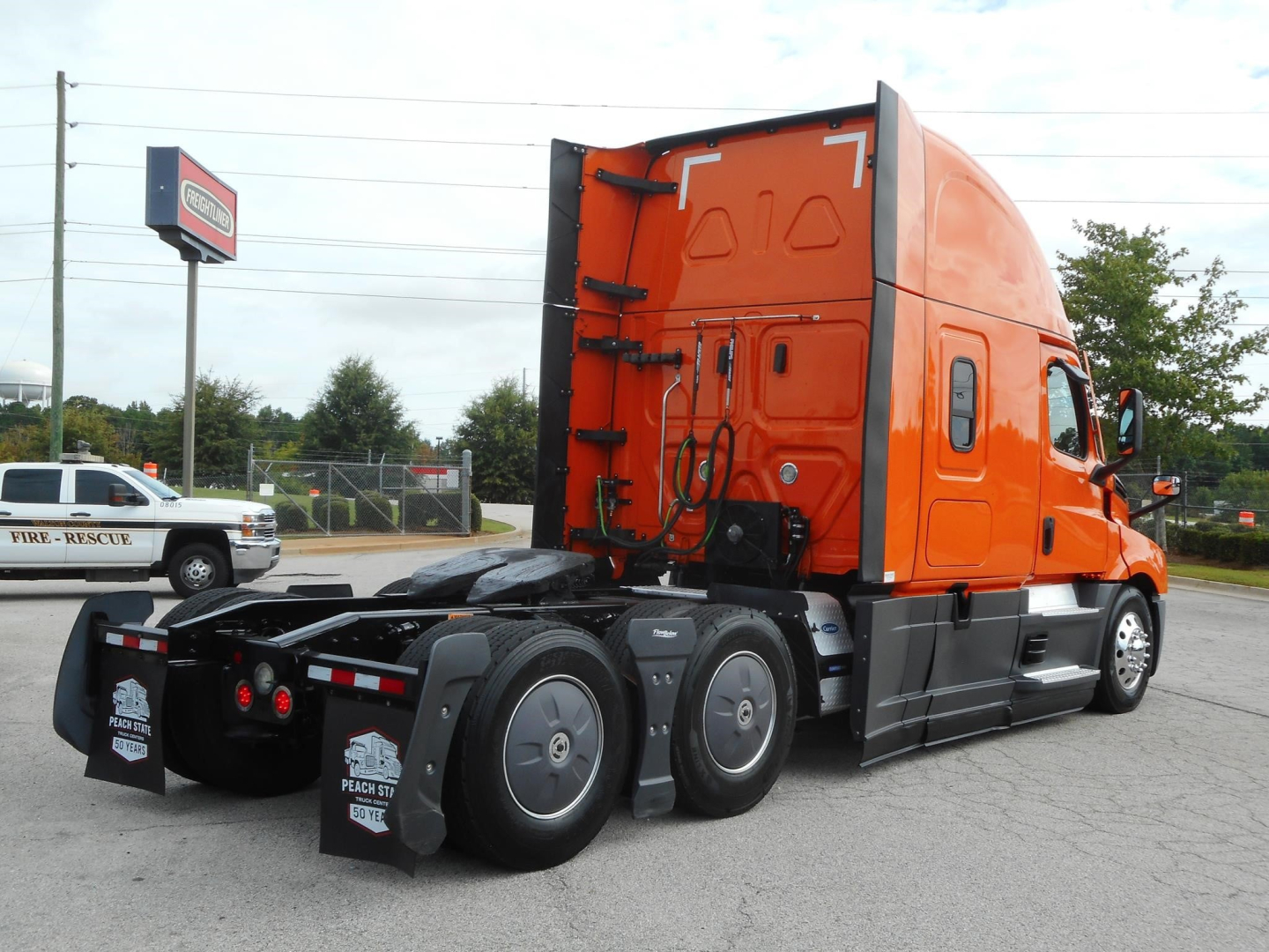 Freightliner Cascadia truck rear side view showing orange sleeper cab and dual rear axles.